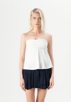Witte strapless top van een textiele stof met een uitlopende zoom, gecombineerd met een donkerblauwe minirok met ruches. Vlakke, gladde ondergrond en zachte draping.