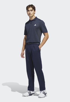 Mand står med den ene hånd i lommen iført navy polo shirt, navy bukser, sort bælte og hvide sneakers med sorte striber.