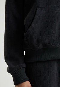 Schwarzes, strukturiertes Sweatshirt aus Stoff mit Tasche, gerippten Bündchen und Saum, sichtbar am Arm und an der Taille einer Person.