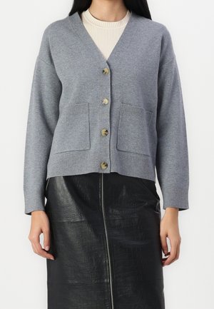 Femme portant un cardigan gris boutonné avec des poches devant et une jupe en cuir noir texturée avec une fermeture éclair sur le devant.