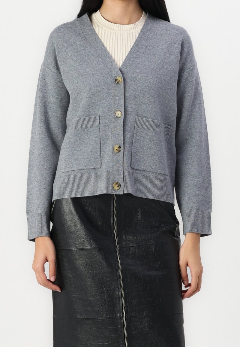 Femme portant un cardigan gris boutonné avec des poches devant et une jupe en cuir noir texturée avec une fermeture éclair sur le devant.