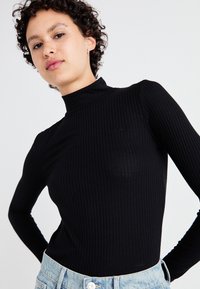 Juodas raštuotas ilgomis rankovėmis suklastotas turtleneck iš minkštos medžiagos, prigludusi forma, minimalūs akcentai; derinamas su šviesiais džinsais.