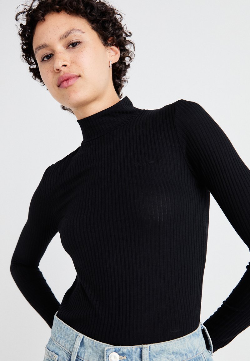 Juodas raštuotas ilgomis rankovėmis suklastotas turtleneck iš minkštos medžiagos, prigludusi forma, minimalūs akcentai; derinamas su šviesiais džinsais.