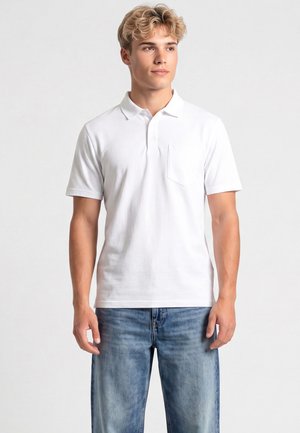 Jeune homme aux cheveux blonds bouclés portant un polo blanc uni à manches courtes et un jean bleu, debout devant un fond clair.