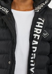 Chaqueta puffer negra con acabado brillante, con cierre de cremallera, secciones grandes acolchadas y detalles de logo impreso en blanco en el forro.