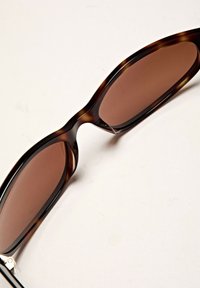 Occhiali da sole tartarugati con lenti marroni, forma aerodinamica e montatura in plastica liscia. Presentano una curvatura sottile e minimi accenti metallici.