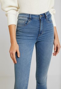 Mujer vistiendo jeans ajustados de color azul claro y un suéter de punto blanco, de pie con las manos relajadas a los lados frente a un fondo liso.