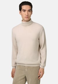 Pull à col montant beige clair avec une texture douce, une coupe décontractée et des poignets et un ourlet côtelés. Porté avec un pantalon en velours côtelé beige.