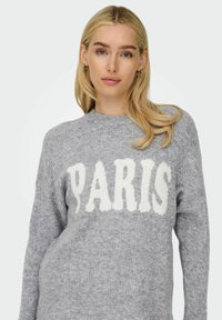 Pull en tricot gris à col rond avec l'inscription "PARIS" en lettres blanches, d'une texture bouclée. Il dispose de manches longues et d'une coupe décontractée.