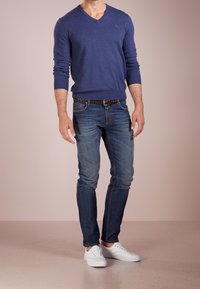 Marinblå V-ringad tröja, slim-fit blå jeans, vita sneakers. Tröjan har långa ärmar; jeansen har lätt slitning och ett brunt vävt bälte.