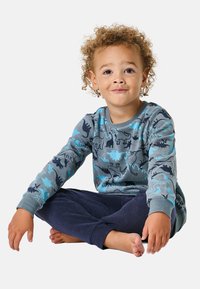 Graues Langarmshirt mit einem Dinosaurierdruck in Blau und Dunkelblau, kombiniert mit dunkelblauen Hosen, sitzendes Kind mit lockigem Haar.