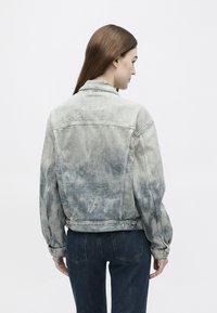 Sieviete ar gariem brūniem matiem, valkājot izbalējušu pelēku džinsa jaku un tumši zilas džinsas, kas stāv ar aizmuguri pret kameru uz balta fona.