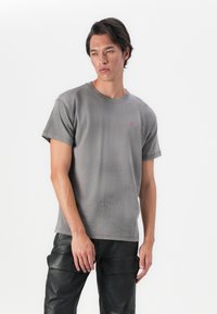 T-shirt cinza em algodão com mangas curtas, apresentando uma textura subtil e um pequeno logótipo vermelho no peito. Combinado com calças cargo pretas.