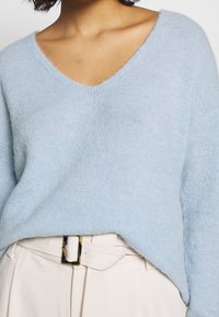 Jasnoniebieski sweter z dekoltem w serek, wykonany z miękkiego, teksturowanego materiału. Charakteryzuje się luźnym krojem z opuszczonymi ramionami i jest zestawiony z beżowymi spodniami z wysokim stanem.