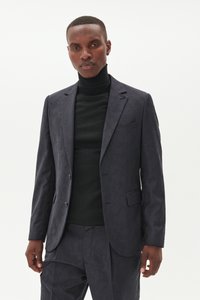 Blazer azul marino con un sutil patrón tonal, diseño de un solo pecho, solapas con muesca y dos botones plateados. Llevado sobre un cuello de tortuga negro.