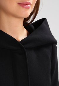 Prenda negra con capucha hecha de tejido suave, que presenta un escote ceñido y una capucha estructurada. Textura lisa sin patrones visibles ni herrajes.