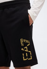 Άτομο που φοράει μαύρο σορτς με χρυσό λογότυπο EA7 Emporio Armani στο αριστερό μηρό, το χέρι βρίσκεται στην πλάγια τσέπη, εμφανίζεται λευκό πουκάμισο με μακριά μανίκια.