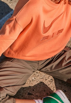 Personne assise en tailleur en plein air, portant un sweat-shirt orange Nike Air, un pantalon beige, des chaussettes blanches et des baskets vertes.