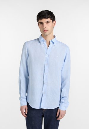 Giovane uomo che indossa una camicia azzurra chiusa con bottoni e maniche lunghe, e jeans blu scuro, in piedi davanti a uno sfondo bianco semplice.