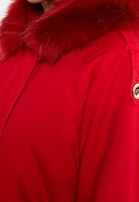 Veste rouge avec un col en fausse fourrure, tissu lisse, fermeture éclair et un bouton rond décoratif sur l'épaule pour plus de détails.