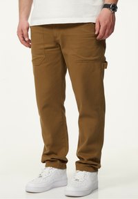 Brown cargo pants made of cotton blend fabric, featuring front patch pockets and a side cargo pocket, paired with white sneakers.
