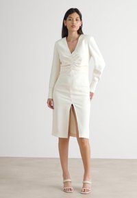 Helles cremefarbenes Midikleid mit langen Ärmeln, V-Ausschnitt, Raffung an der Vorderseite und einem hohen Schlitz bis zum Oberschenkel. Glatte Textur mit minimalen Details.