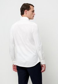 Camicia bianca a maniche lunghe con vestibilità rilassata, colletto a punta e polsini con bottoni. Tessuto liscio con una leggera lucentezza e orlo arrotondato.