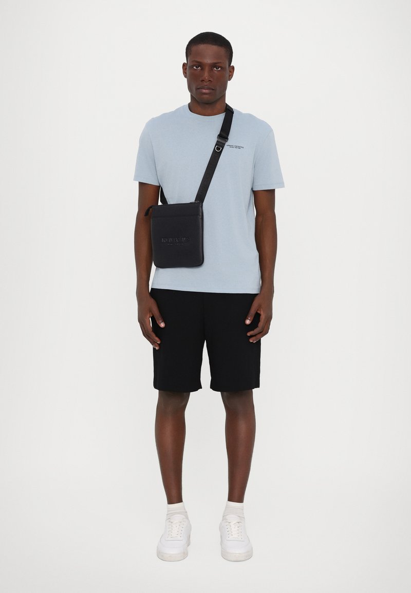 Svart axelväska med präglad text, platt design, rektangulär form, bärs över en ljusblå skjorta, ihop med svarta shorts och vita sneakers.