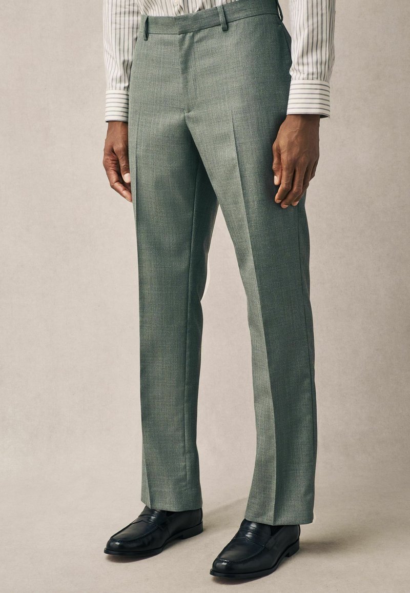 Pantalones ajustados verde claro con una tela texturizada, frente plano y dos bolsillos laterales, combinados con mocasines negros.