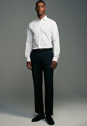 Mann steht und trägt ein weißes Hemd mit Knöpfen, schwarzen Gürtel, schwarze Anzughose und schwarze Anzugsschuhe vor einem schlichten grauen Hintergrund.