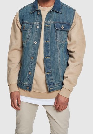 Man draagt een blauwe denimvest over een beige trui en lichtgekleurde broek tegen een egale lichte achtergrond.