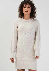 Robe pull en beige clair, avec un col rond, de longues manches ballon, deux poches avant et des motifs découpés en chevrons.