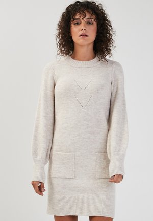 Robe pull en beige clair, avec un col rond, de longues manches ballon, deux poches avant et des motifs découpés en chevrons.