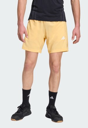 Hombre que lleva pantalones cortos deportivos amarillos, calcetines deportivos negros y zapatillas de correr negras, de pie frente a un fondo liso.