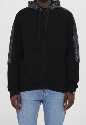 Sweat à capuche noir à fermeture éclair avec des bandes sombres imprimées sur les manches et la capuche, porté par une personne en jean bleu clair sur un fond uni.