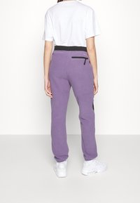 Lila sweatpants med svart midjeband, med en zip-ficka på baksidan och strukturerad tyg. Bärs med en vit T-shirt och vita sneakers.