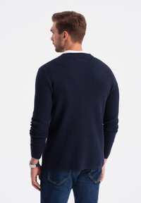 Suéter de punto azul marino con un acabado texturizado y cuello de canalé, combinado con jeans de mezclilla y un reloj plateado en la muñeca.