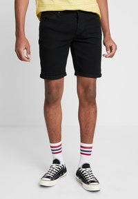 Pantalones cortos de mezclilla negra con dobladillos enrollados, con bolsillos. Combinados con zapatillas negras y calcetines blancos que muestran rayas rojas y azules.