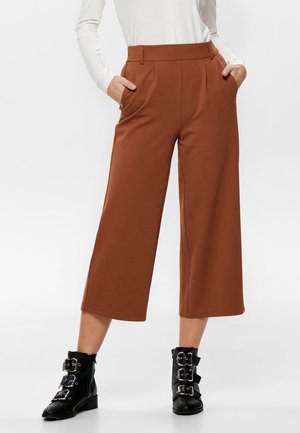 Vrouw draagt hoge taille, wijde cropped bruine broek met handen in de zakken en zwarte enkellaarsjes met gesp.