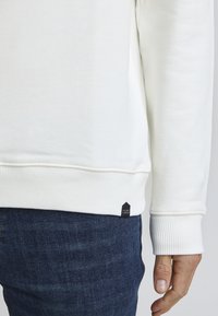 Felpa bianca con polsini e orlo a costine, caratterizzata da un piccolo etichetta nera sul lato inferiore. Indossata con jeans in denim scuro.