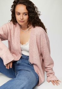 Różowy dzianinowy kardigan o prążkowanej strukturze, otwarty z przodu, z długimi rękawami. Nosi się go na białą bluzkę z koronkowym wykończeniem oraz niebieskie dżinsy z wysokim stanem.