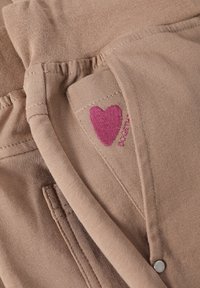 Beżowa tkanina o gładkiej fakturze, zdobiona różowym haftowanym sercem i detalami z guzików. Projekt zawiera małą kieszeń oraz przeszycia dekoracyjne.