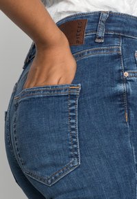 Jeans de mezclilla azul oscuro con un ajuste de tiro medio, parche de cuero marrón en la cinturilla y detalles de costura visibles. El bolsillo es parcialmente visible.