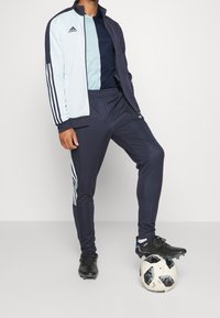 Chándal Adidas que presenta una chaqueta azul claro y marino con tres rayas blancas, combinada con pantalones marinos a juego. Calzado de fútbol negro y un balón decorado.