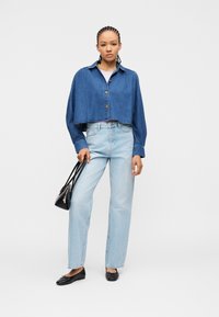 Γυναίκα με πλεξούδες που φοράει μια κοντή μπλε τζιν ζακέτα, ανοιχτό μπλε τζιν παντελόνι, μαύρα φλατ παπούτσια, κρατώντας μια μικρή μαύρη τσάντα.