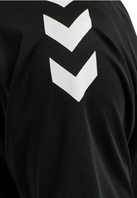 Zwarte sporttrui met een gladde textuur, met drie witte chevron-strepen die diagonaal op het bovenste gedeelte zijn geplaatst.