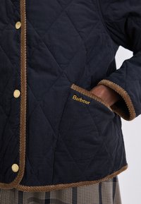 Casaco acolchoado azul-marinho com acabamento em corduroy castanho e botão de pressão dourado; possui um bolso lateral com o logótipo "Barbour" em costura amarela.