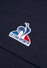 Un tessuto blu navy presenta un logo ricamato con un gallo stilizzato, accenti rossi e blu, e "LE COQ SPORTIF" in testo bianco.