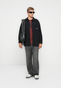 Svart skjorte med knapper og "GAP" logo, vinrød t-skjorte med "A" trykk, grå jeans, svarte sko og en svart skinn skulderveske.