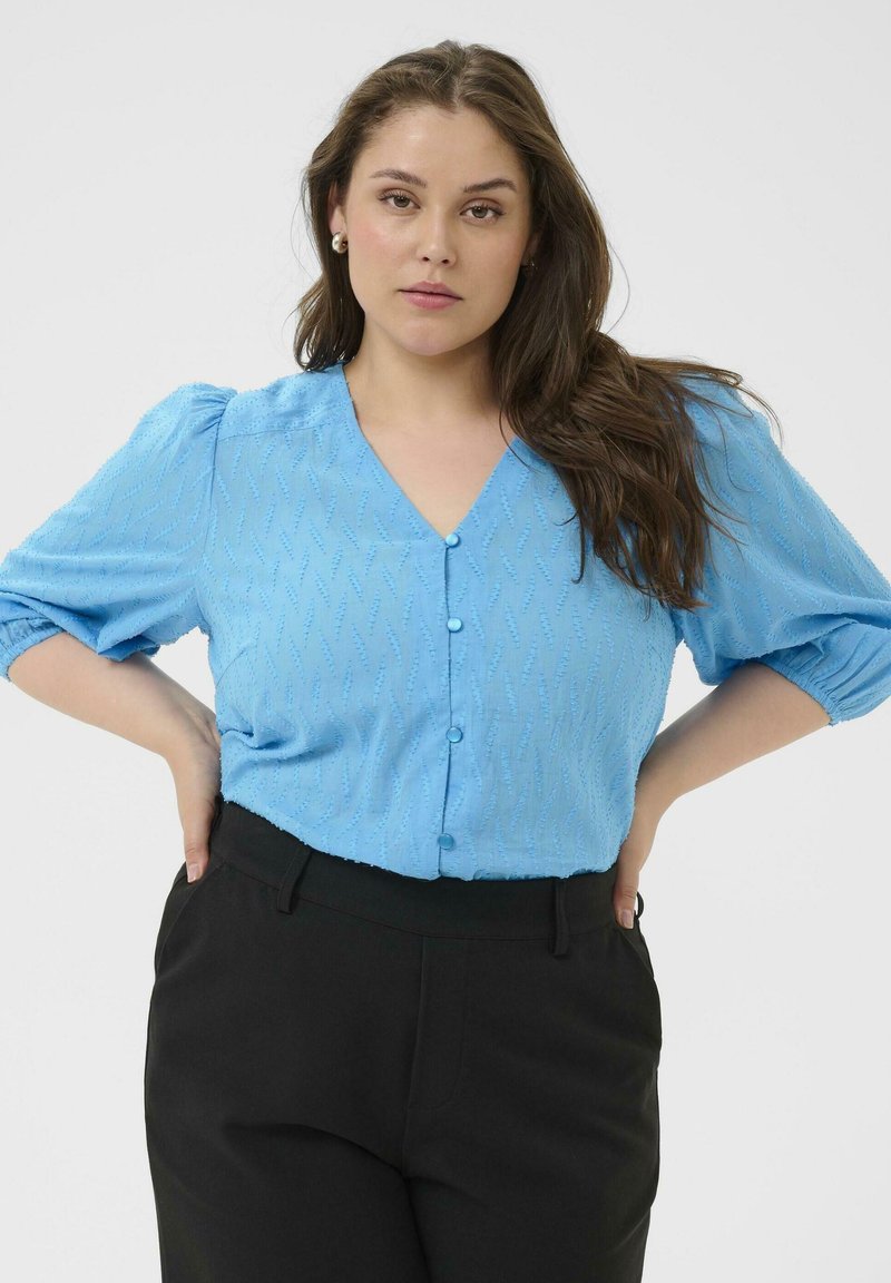 Blouse bleu clair avec un motif texturé, col en V, et manches courtes bouffantes, associée à un pantalon noir. Des boutons ornent le devant.